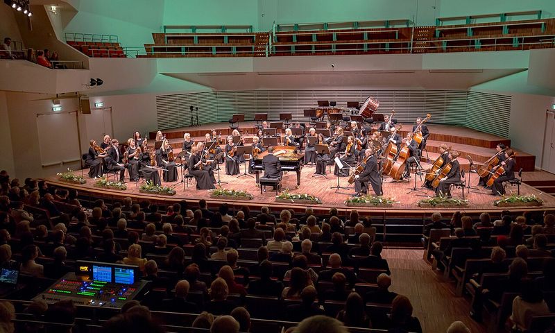 Orchestră simfonică și pian pe scena unei săli de concert, în fața publicului